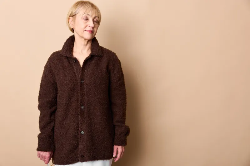 Elderly woman portrait in a studio, wearing a brown jacket and white shirt, beige background, calm smile, relaxed posture, soft light, warmth, wellbeing, gentle care