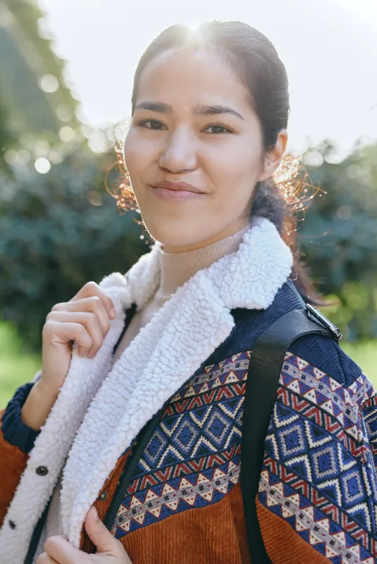 Smiling woman outdoors wearing a patterned jacket and backpack, enjoying sunny weather and nature, casual fashion and travel vibe with warm light and relaxed confidence