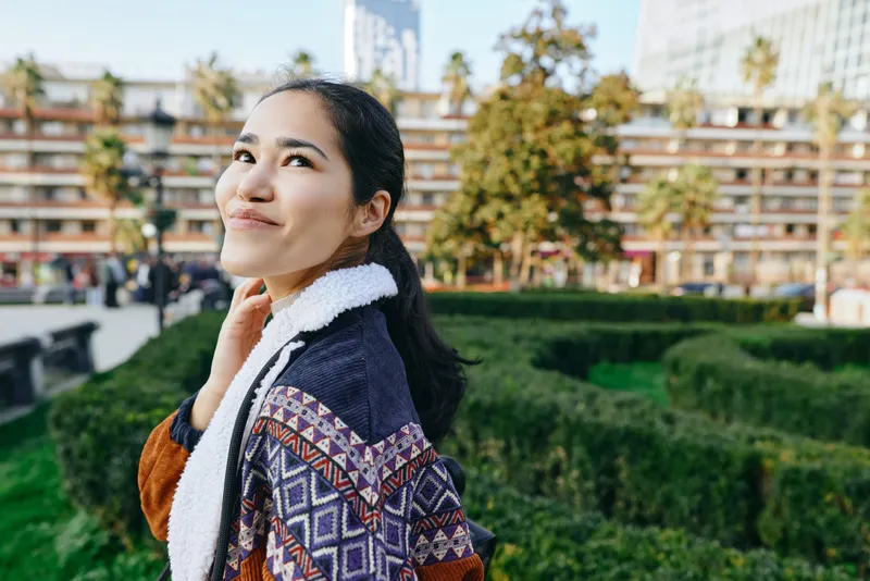 Woman in a patterned shawl outdoors in a sunny park, gazing upward with a gentle smile, capturing a moment of calm, contemplation, and urban nature blending with greenery