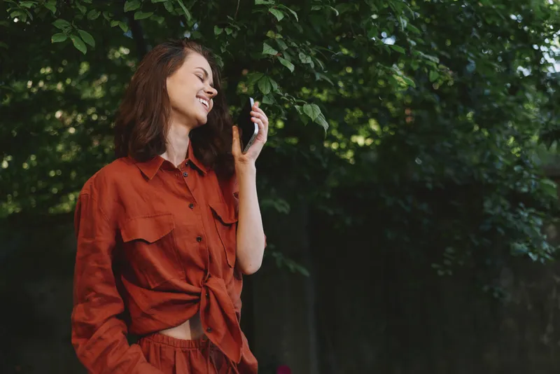 Cheerful woman outdoors, smiling while talking on a phone, red outfit, sunlit greenery background, relaxed lifestyle moment, natural colors, airy atmosphere