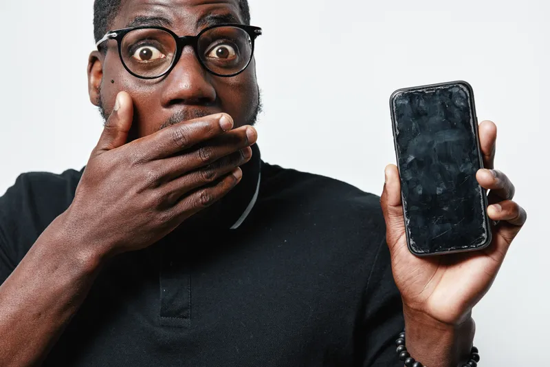 African American man with dark skin and glasses showing shocked expression covering mouth holding smartphone with blank screen isolated on white background.
