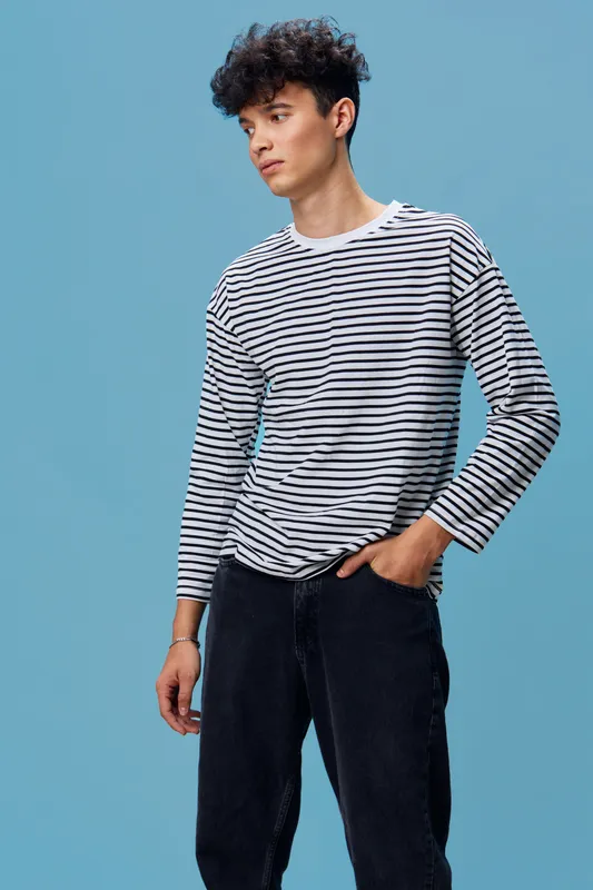 Young man wearing striped long sleeve shirt and dark pants posing against blue background, looking thoughtful with curly hair in casual fashion style.