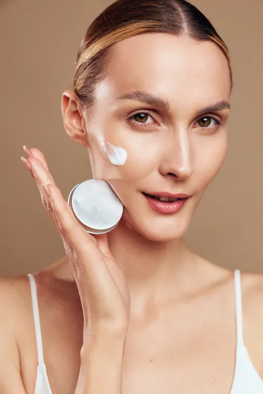 A young woman with clear skin applies white facial cream on her cheek while holding an open jar. The close-up shows skincare routine and beauty care concept with a neutral background.