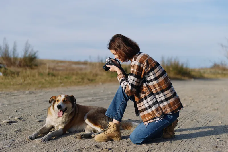 Photographer kneeling in a sandy outdoor setting, capturing a playful dog with a compact camera, casual plaid jacket, and boots, sunlight and wide open space for a relaxed nature moment
