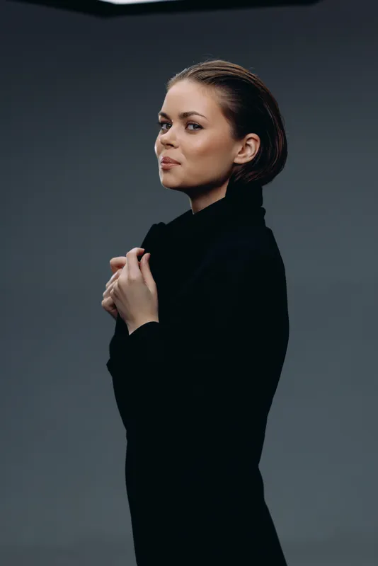 Young woman in black coat posing confidently against dark background with subtle lighting