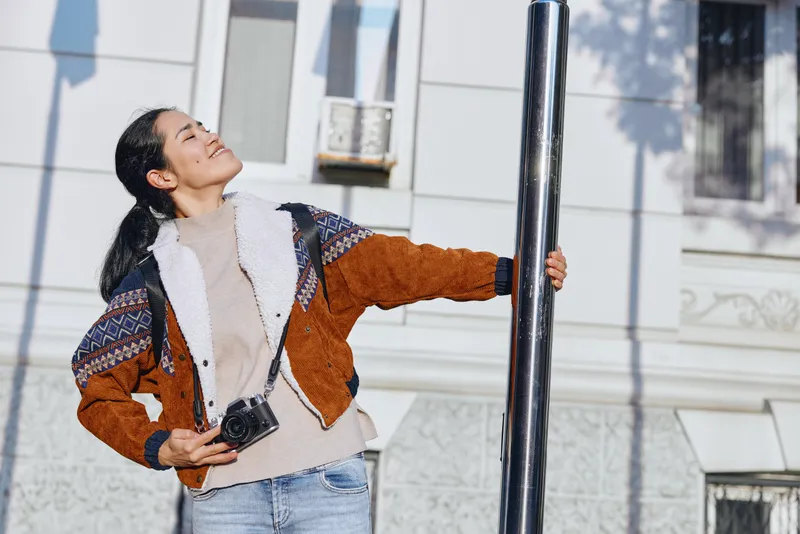 Young woman holds a camera and reaches toward a street lamp, outdoors, wearing a colorblock jacket and denim, capturing a candid urban moment with confident pose