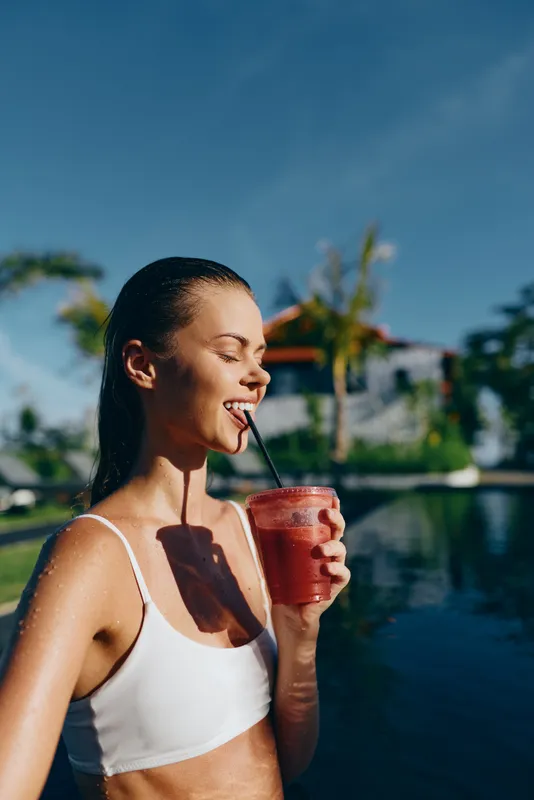 A young woman in a white swimsuit enjoys a refreshing red smoothie by the pool under bright blue sky, embodying a relaxed and healthy lifestyle moment outdoors.