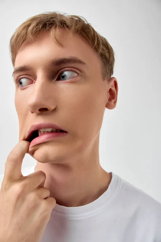 Close up of a young man with a pensive expression, biting his lip and glancing sideways, conveying anxiety and uncertainty, against a plain white background, casual white tee, intimate face detail,