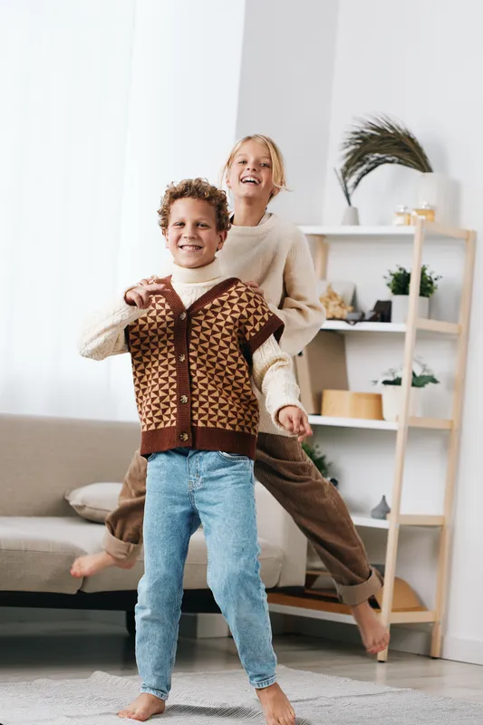 Authentic scene of playful siblings in a cozy home, two children smiling and dancing while wearing casual knitwear. A light, wholesome moment capturing everyday school life and lively lifestyle.