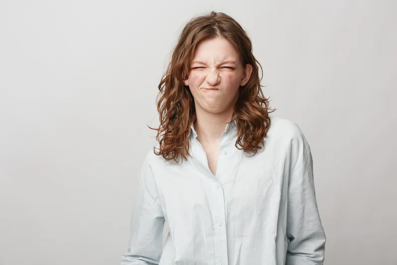 Young woman with curly hair wearing white shirt makes funny grimace with eyes closed and lips pressed. Isolated on light background. Concept of emotions, humor, and playful mood.