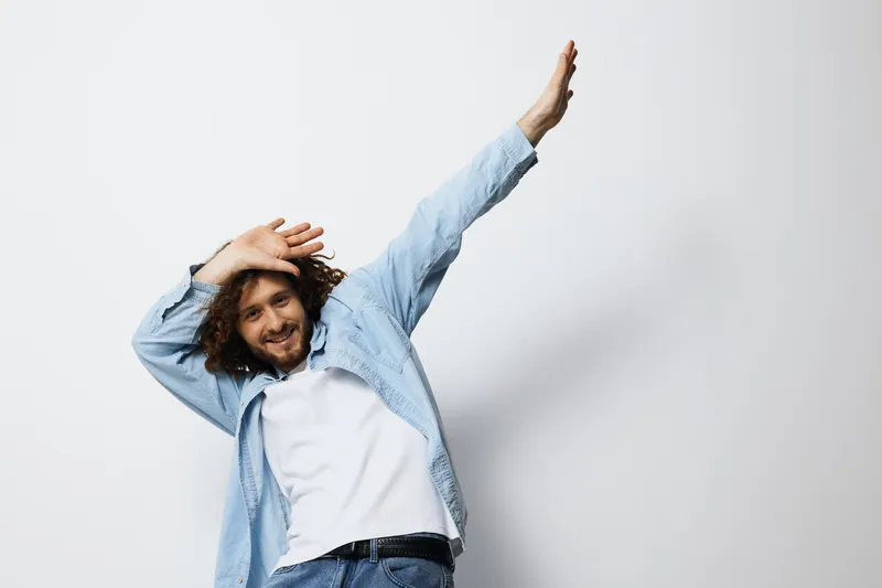A joyful young man with curly hair in a light blue shirt poses playfully against a white background, expressing happiness and energy in a casual style