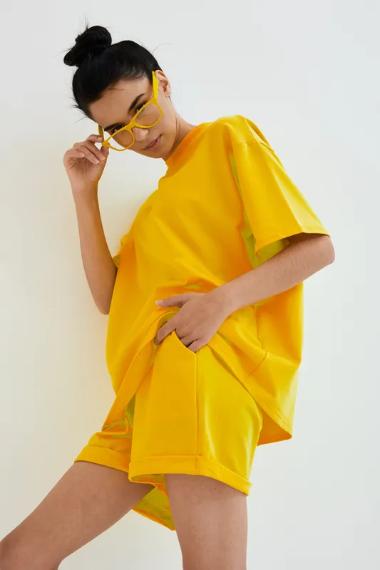Woman in bright yellow casual outfit and glasses posing indoors against white background, showcasing relaxed and modern style with minimalistic composition.