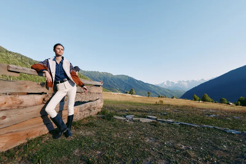 Man in cowboy boots leans on wooden fence in mountain meadow landscape, outdoors and rural scene with rolling hills, clear sky and solitary traveler enjoying nature and freedom