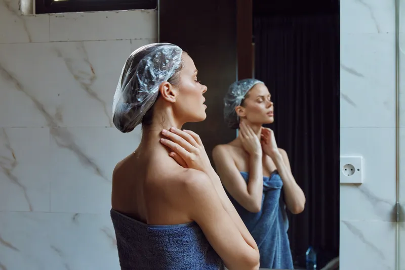 Woman with shower cap wrapped in towel touching neck and looking in bathroom mirror, natural skincare routine, fresh clean beauty.