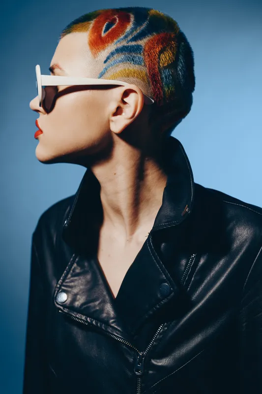 Woman with colorful shaved hair wearing white sunglasses and a black leather jacket, posing in profile against a blue background, showing trendy and edgy style.