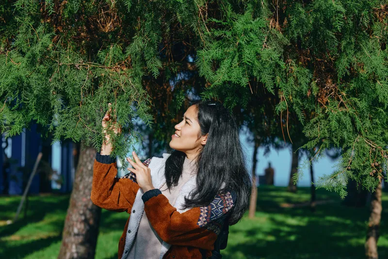 A woman outdoors admires leaves on a tree, reaching gently to interact with greenery in a sunlit garden setting, capturing a moment of calm, curiosity, and connection with nature.