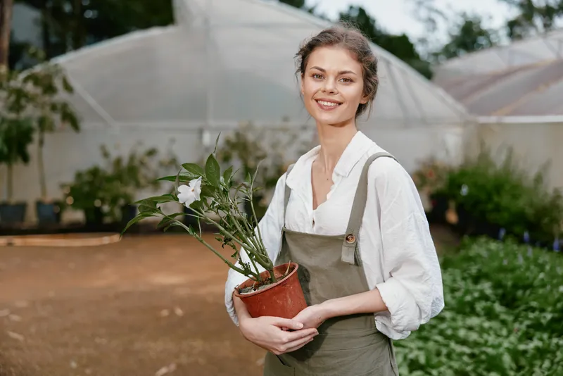 Young woman in green apron smiling with a potted plant in a garden nursery surrounded by greenery, perfect for conveying healthy living and gardening concepts
