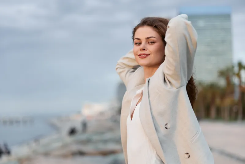 Confident woman enjoying outdoor urban lifestyle with casual coat and relaxed smile in soft natural light, city and waterfront background, warm tones