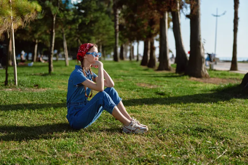 Lifestyle woman wearing modern boho-western denim street style outfit sitting on grass in sunny park, thoughtful mood, film color tones enhancing natural scene.
