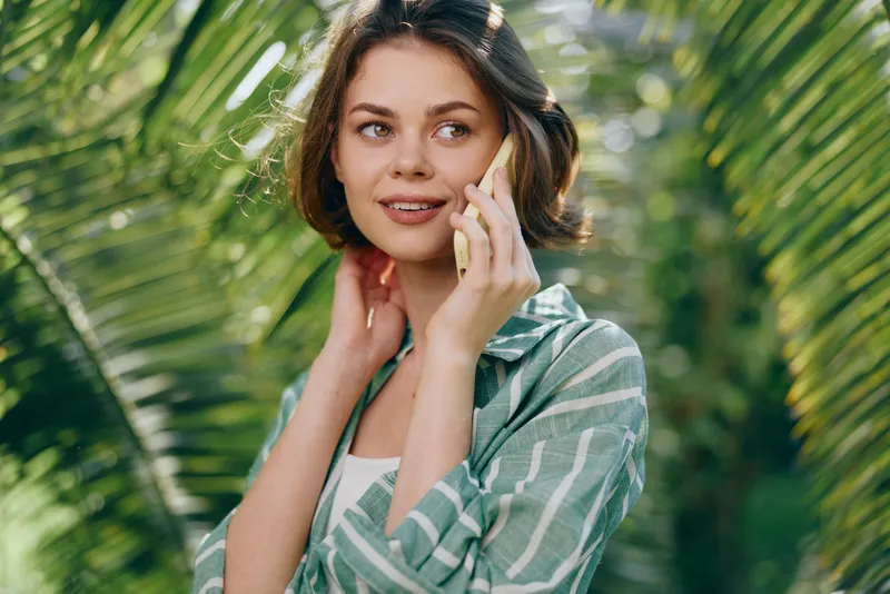 A woman smoothly talks on a smartphone outdoors, standing among lush palm leaves in bright natural light, wearing a green striped shirt.