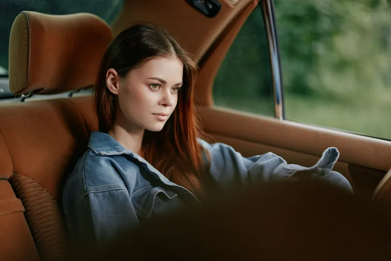 A contemplative young woman in denim attire sits in a vintage car, gazing thoughtfully outside, surrounded by greenery on a serene day.