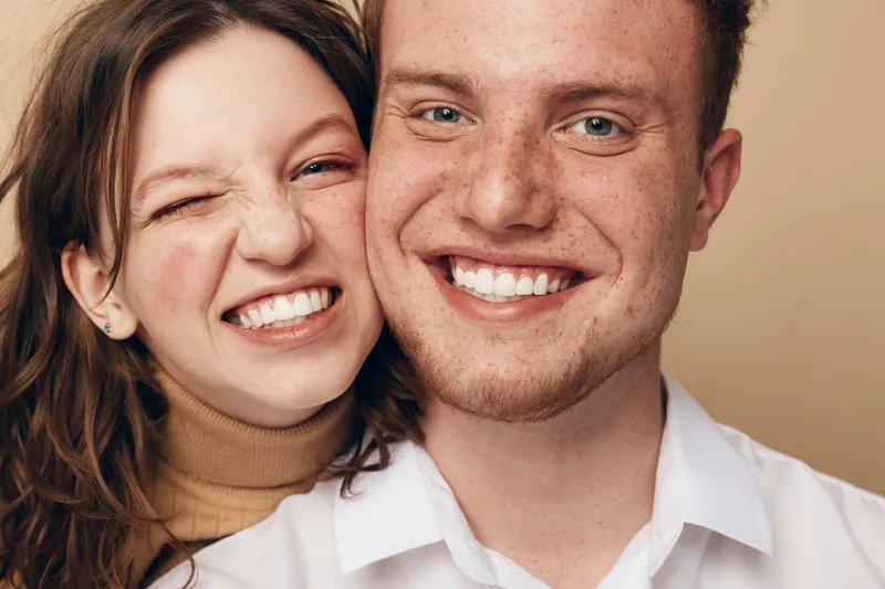 Smiling young couple with freckles and casual clothes pose close together on beige background. Happy man and woman show genuine joy and affection in warm natural light.