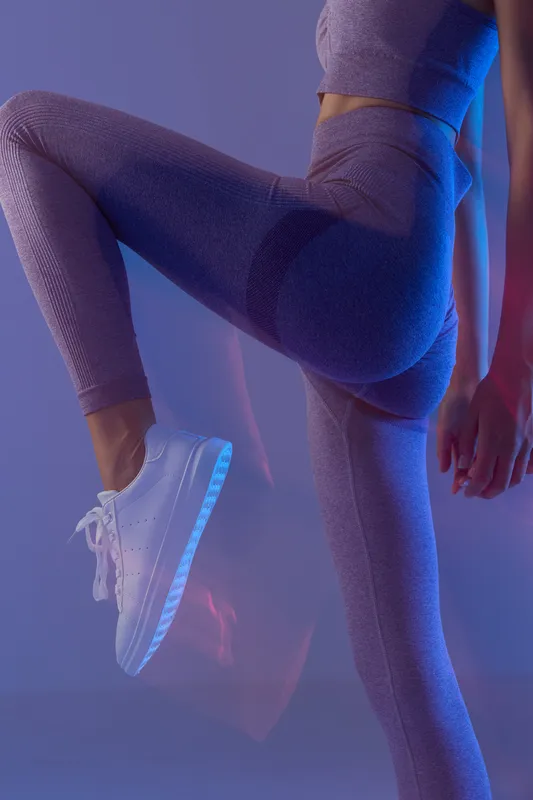 woman sport fitness wearing purple leggings and white sneakers, active pose focusing on leg muscles and dynamic movement in colored lighting