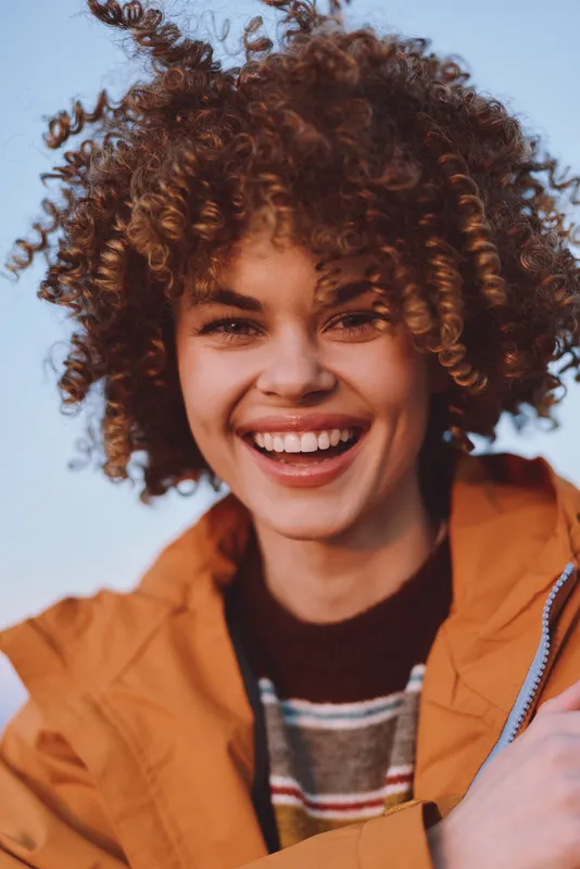 Smiling woman with curly hair, closeup portrait in a casual orange jacket, outdoors in soft daylight, warm lifestyle feeling, wind lifts curls as she laughs, confidence and self care