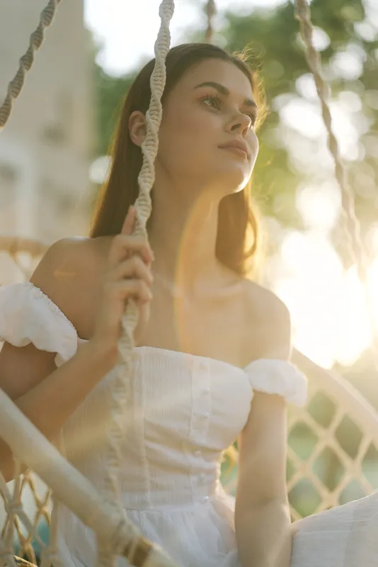 Young woman in a white dress sitting on a swing, enjoying a warm sunset, radiating calm and peaceful emotions, surrounded by a natural green background
