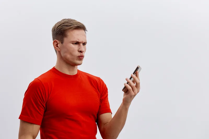 Man wearing red t-shirt looking confused at smartphone isolated on plain white background studio portrait. People lifestyle concept