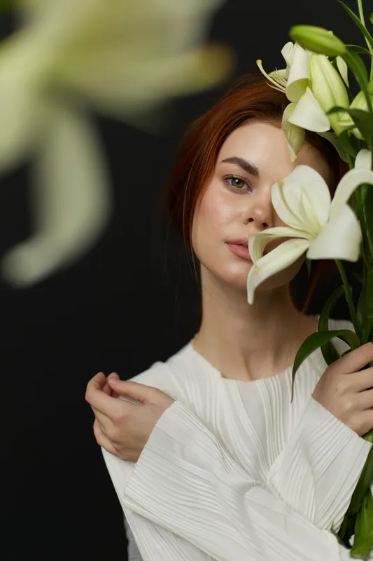 Young woman with long red hair in a white blouse, holding white lilies against a dark background, conveying elegance and serenity