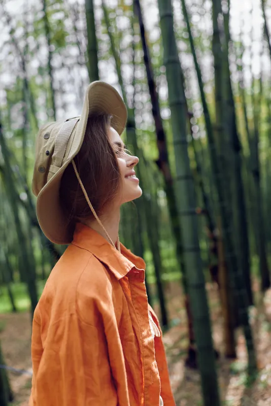 Woman profile smiling in bamboo forest wearing wide brim hat and orange shirt, portrait of nature lover enjoying an outdoor walk in a peaceful green grove and relaxed mood.