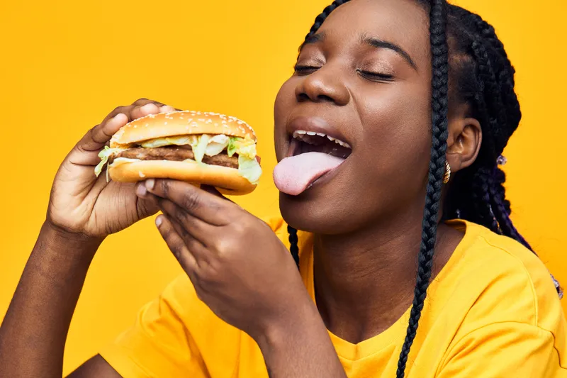 Young Black woman joyfully savoring a delicious hamburger, wearing a bright yellow shirt against a vibrant yellow background