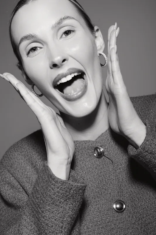 Happy Caucasian woman with light skin and open mouth poses cheerfully in warm knitted sweater with earrings in monochrome studio portrait. Concept of joy and emotion.