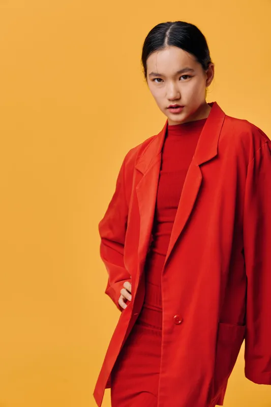 Woman wearing red coat and dress posing with hand on hip against yellow background. Portrait of confident fashion model with sleek hairstyle and serious expression in studio lighting.