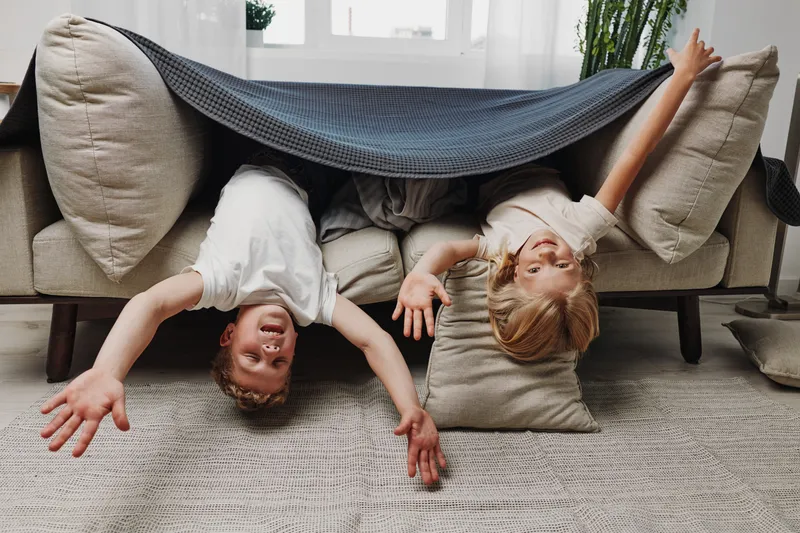 Two happy children upside down under a couch with a blanket, playing indoors on a carpet. Concept of childhood joy, imagination, fun, and family moments at home.