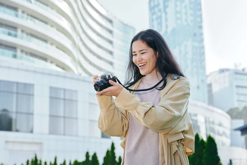 Camera wielding woman enjoys outdoor urban photography in daylight, capturing candid moments with a joyful smile amid modern city architecture and glass towers today.