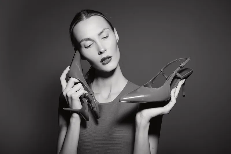 Slim white woman with pale skin holds high heel shoes near face, eyes closed in black studio. Concept of fashion, elegance, footwear and beauty portrait.