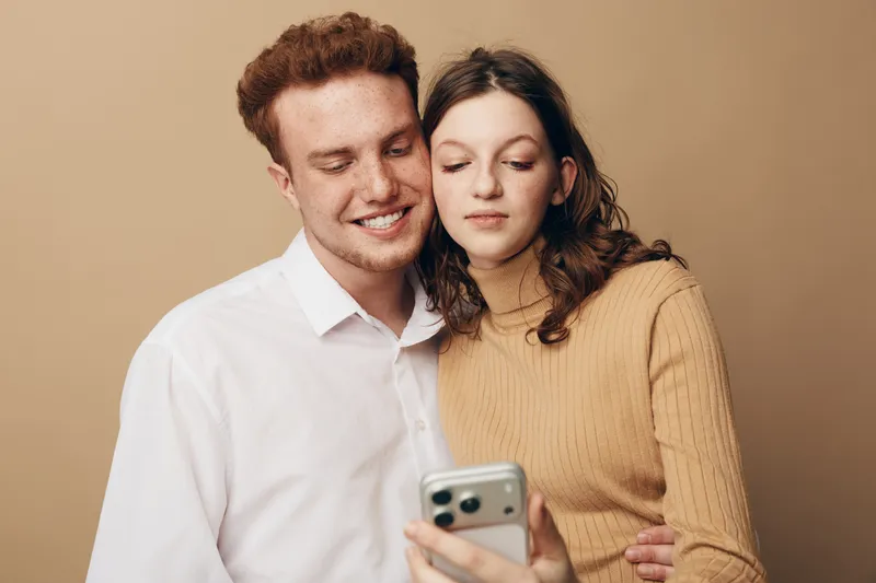 Young couple with light skin and brown hair looks at smartphone together, smiling and relaxed. Casual clothing and neutral background create warm, intimate atmosphere of shared moment.