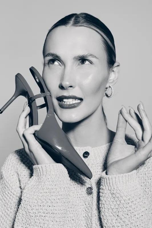 Young Caucasian woman with light skin holds stylish high heel shoe near face, posing with thoughtful expression in studio. Concept of fashion and style.