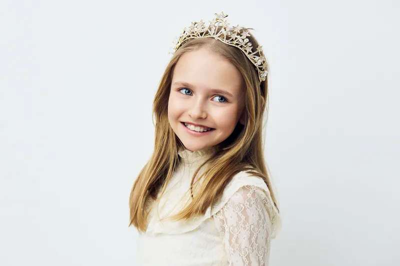 Young girl with a joyful expression wearing a beautiful crown and elegant dress against a soft white background, perfect for children s portraits or princess themes