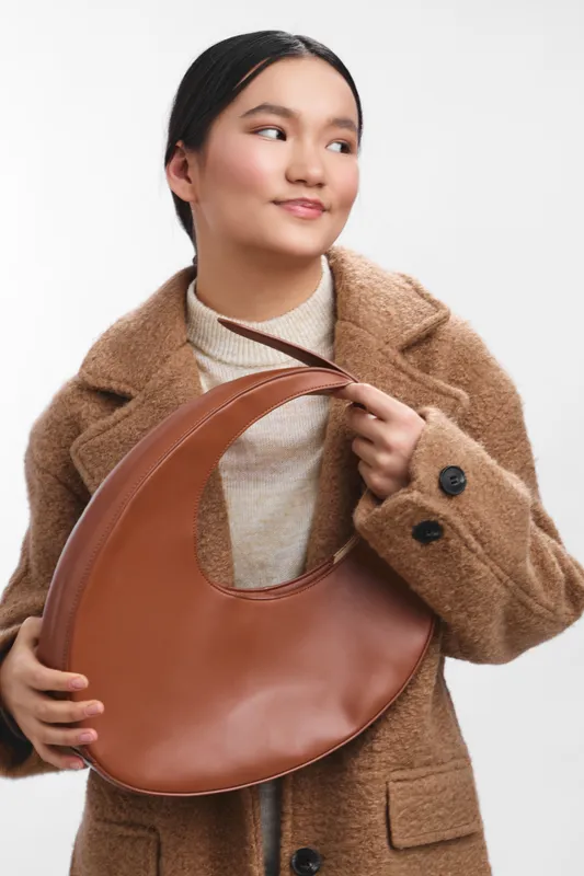 Asian woman poses with a large brown handbag, wearing a beige turtleneck and brown coat. She stands against a light backdrop, exuding style and confidence while showcasing fashion and accessories.
