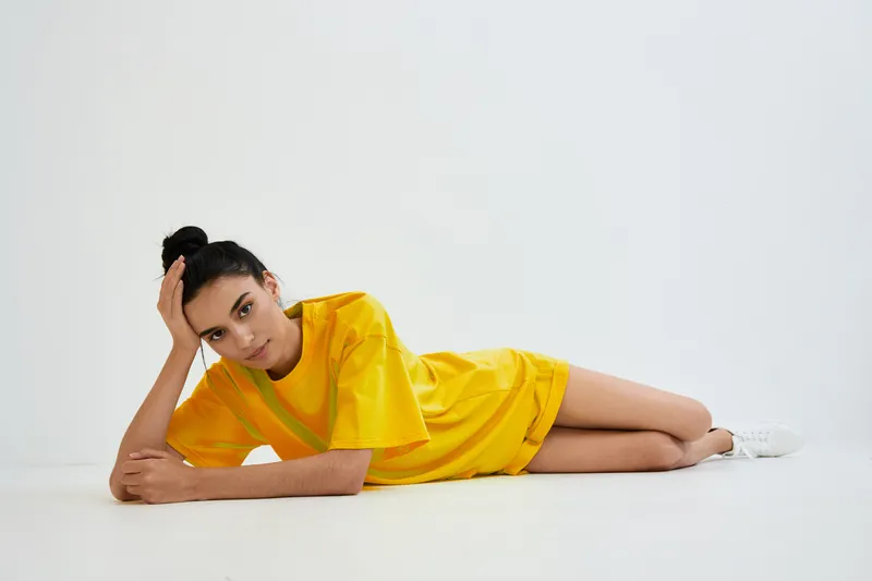 Young woman lying down in a yellow casual dress and white sneakers, posing thoughtfully against a plain white background, with relaxed and calm expression.