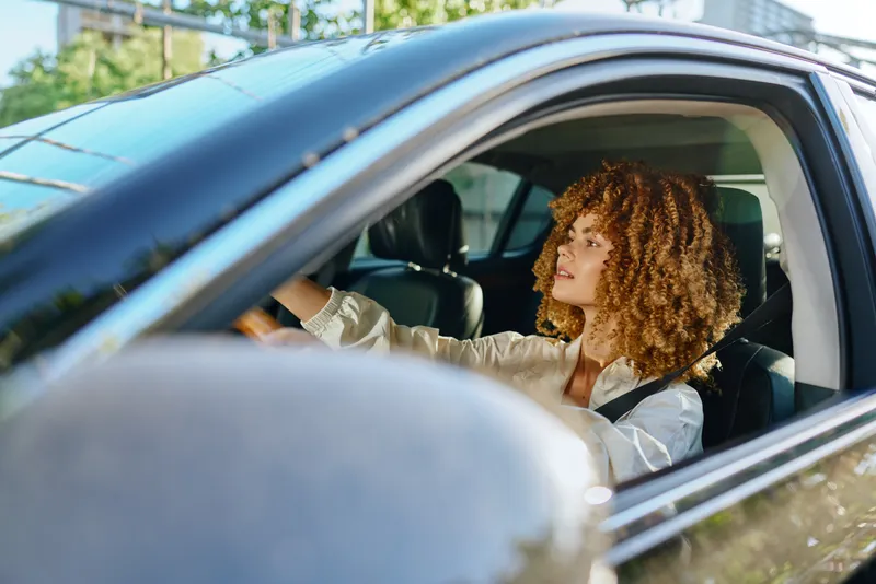 Young woman driving car with curly hair, wearing seatbelt and white shirt, focused on city street, bright daylight, safe travel concept.