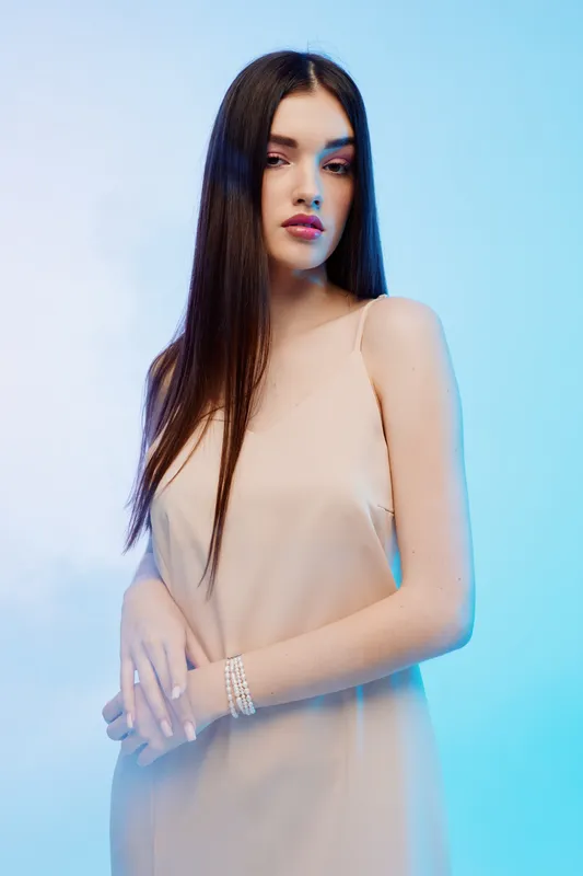 Young woman with long dark hair wearing a beige dress poses elegantly against a soft blue background. Concept for beauty, fashion, and style with a calm mood.