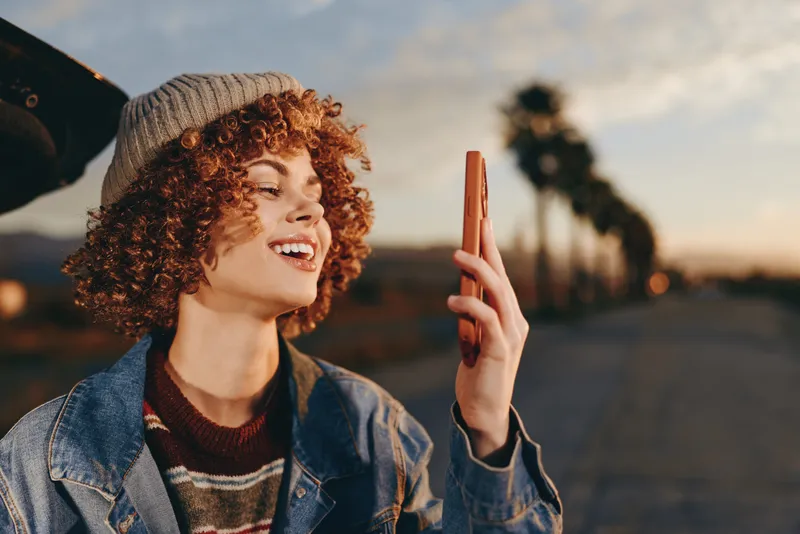 Woman outdoors, smiling with curly hair, denim jacket and knit beanie, holding smartphone and chatting, candid sunset road background, warm golden light, casual lifestyle moment