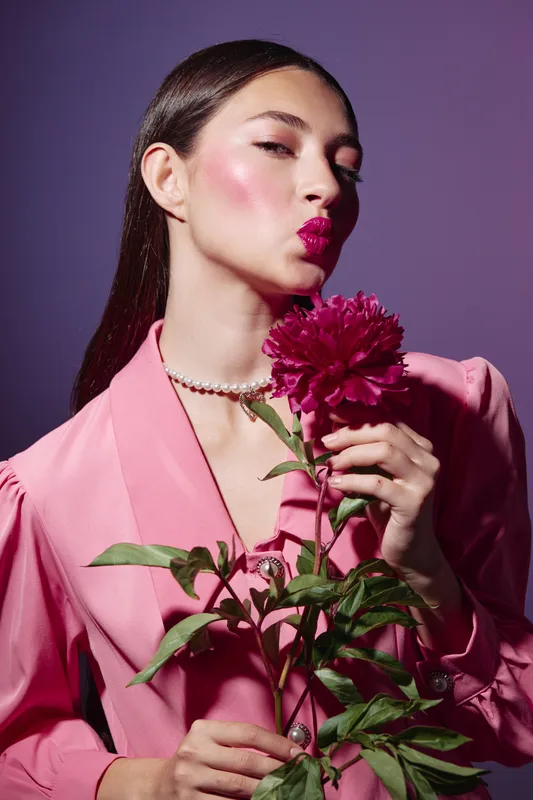 Young woman with glossy makeup puckering lips and holding bright pink peony. She wears a peach blazer and pearl necklace against purple background. Concept for beauty creative and fashion.