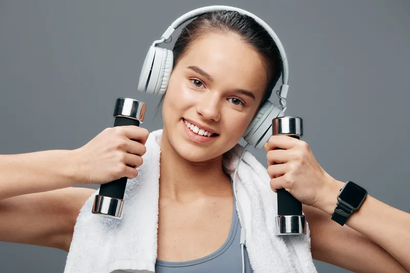 Athletic woman with towel over shoulders, lifting dumbbells and wearing headphones during a focused gym workout, showcasing strength, endurance and motivation in a modern fitness setting.
