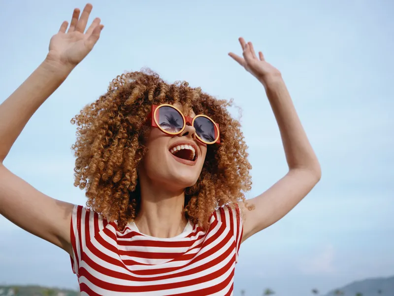 Joyful person outdoors wearing a striped shirt and round sunglasses, raising arms in celebration under a clear blue sky, radiating summer happiness and carefree energy.