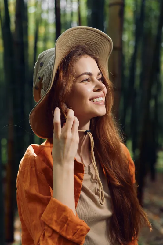 Woman portrait smile in a sunlit forest wearing a wide brim hat and casual outfit, long hair and relaxed expression. Natural outdoors scene with warm sunlight and peaceful nature mood.